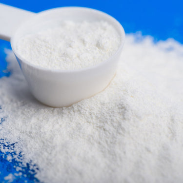 A plastic scoop filled with white powder sitting in a mound of Maxi-BCAA powder, on a blue background